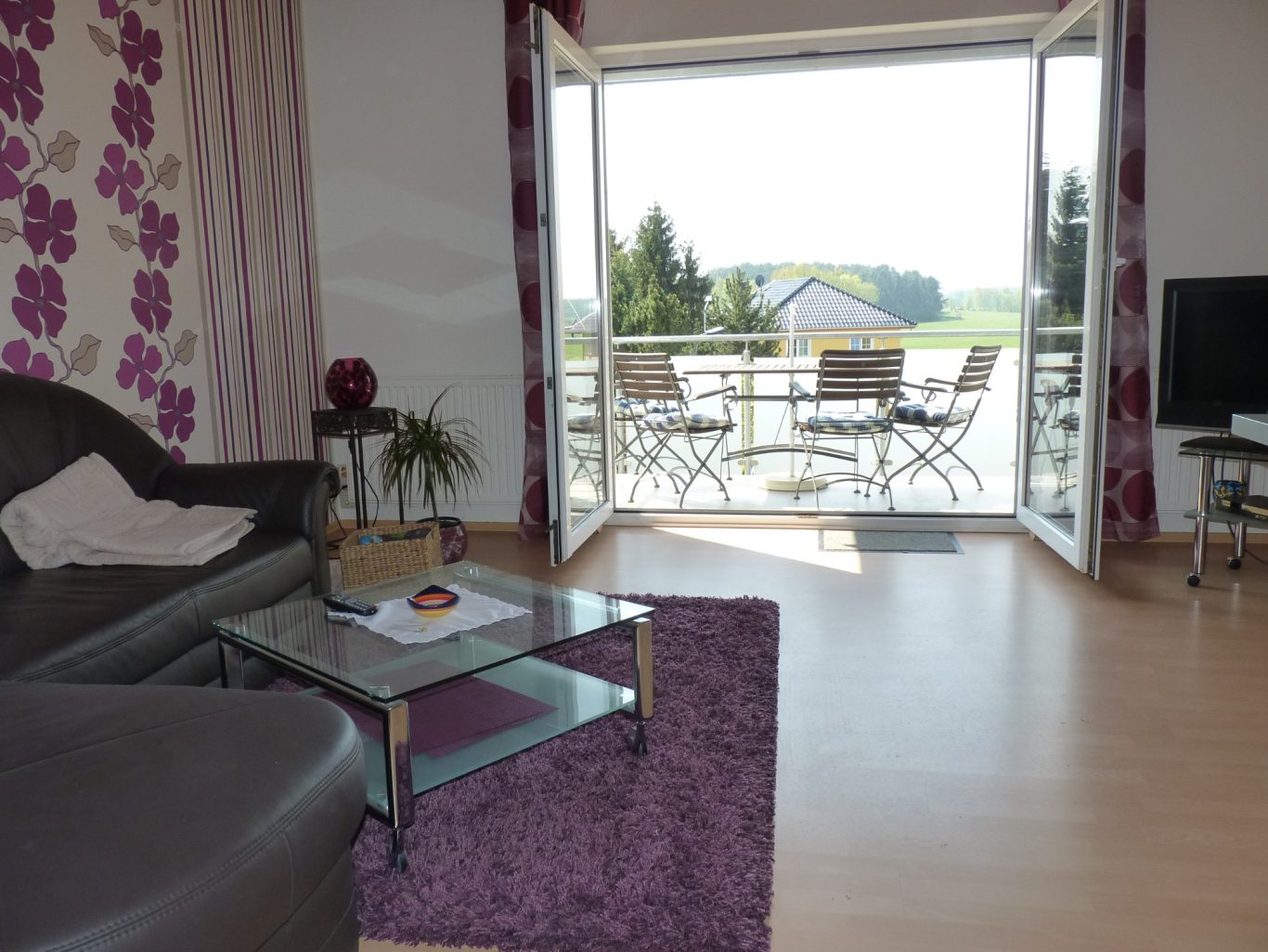 Wiesenblick aus dem Wohnzimmer Wohnzimmer mit Sofa, Glastisch und Blick auf Terrasse mit Stühlen und grüner Landschaft.