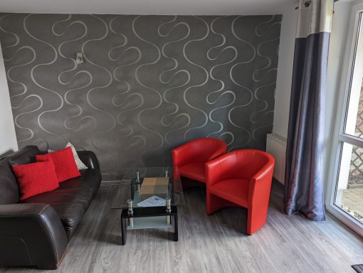 Sitzecke Ferienwohnung Wohnzimmer mit grauer Wand, braunem Sofa und zwei roten Stühlen. Holztisch mit Zeitschriften.