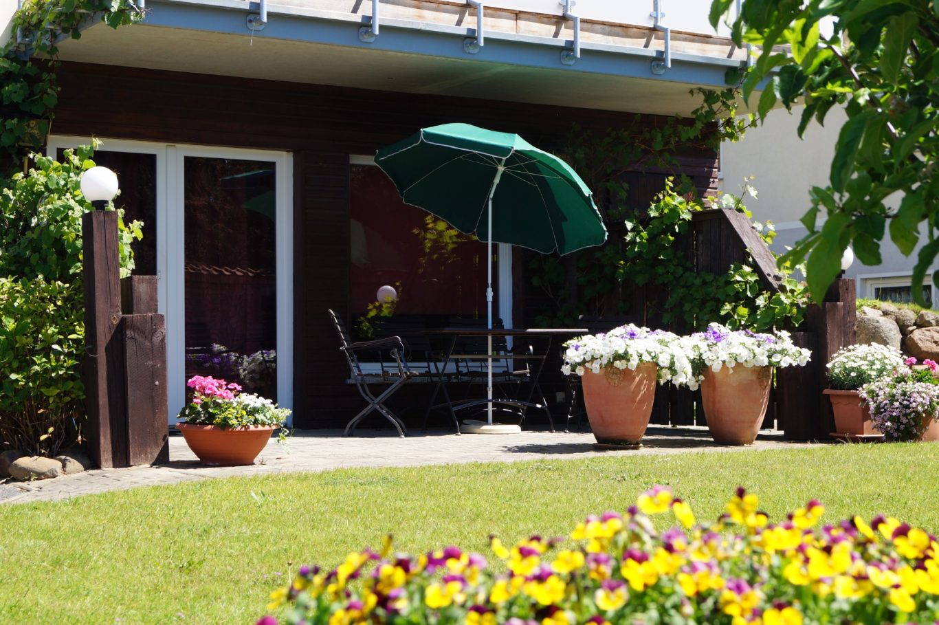 Terrasse Gartenapartment Terrasse mit grüner Sonnenschirm, Blumenbeeten und gemütlichen Sitzgelegenheiten.
