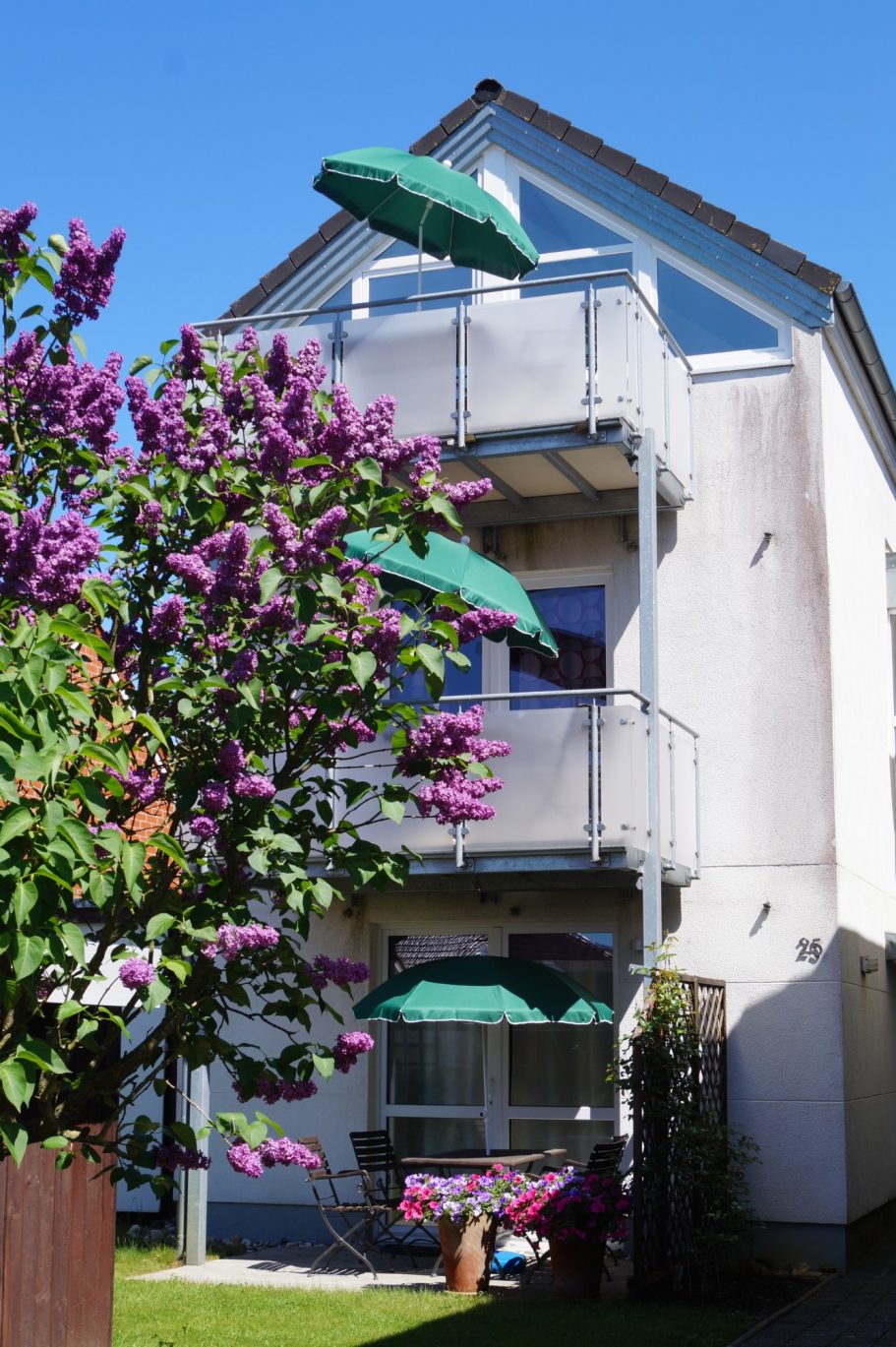 Gästehaus Haus mit Balkon und grünen Markisen, umgeben von lila blühenden Pflanzen.